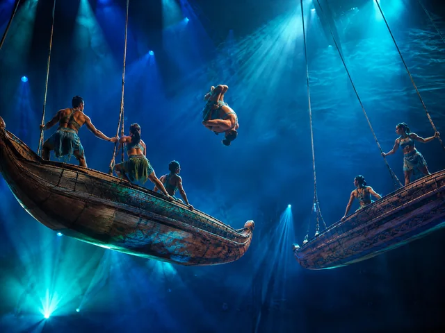 Acrobats flying from swings shaped like a voyaging canoe under blue stage light